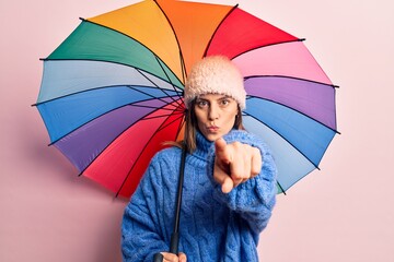 Young beautiful woman holding colorful umbrella pointing with finger to the camera and to you, confident gesture looking serious