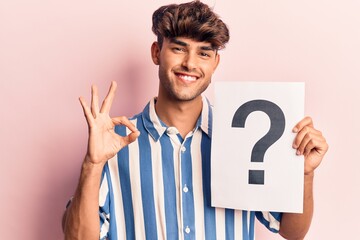 Young hispanic man holding question mark doing ok sign with fingers, smiling friendly gesturing excellent symbol