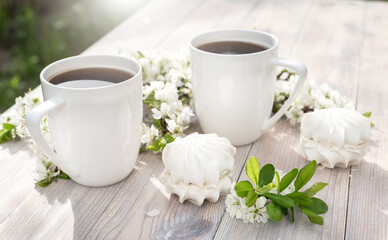 Two beautiful porcelain coffee cups with marshmallow dessert decorated with white cherry flowers on wooden table in spring garden in setting sun light.