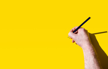 Male hand about to write on blank yellow background with black pencil
