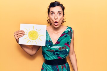 Young beautiful hispanic woman holding sun draw scared and amazed with open mouth for surprise,...
