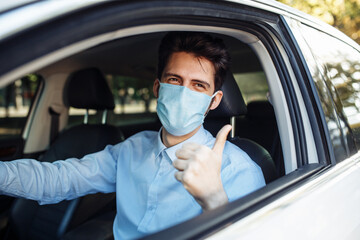 Fototapeta na wymiar Young taxi driver shows thumb up sign sitting in the car and wearing protective sterile medical mask, works hard during coronavirus outbreak. Social distance, virus spread prevention, treat concept.