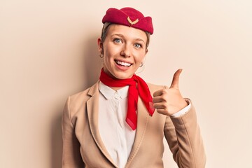 Young beautiful blonde girl wearing stewardess uniform smiling happy and positive, thumb up doing excellent and approval sign