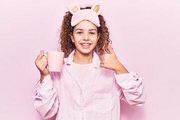 Beautiful kid girl with curly hair wearing sleep mask and pajamas holding coffee smiling happy and positive, thumb up doing excellent and approval sign