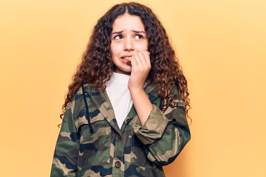 Beautiful kid girl with curly hair wearing camouflage jacket looking stressed and nervous with hands on mouth biting nails. anxiety problem.