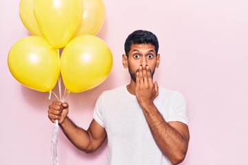 Young latin man holding balloons covering mouth with hand, shocked and afraid for mistake....