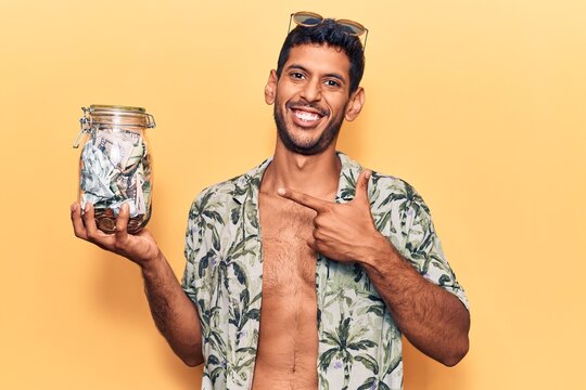 Young latin man wearing summer shirt holding jar of savings smiling happy pointing with hand and finger