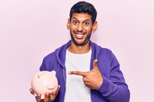 Young latin man holding piggy bank smiling happy pointing with hand and finger