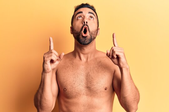Young hispanic man standing shirtless amazed and surprised looking up and pointing with fingers and raised arms.