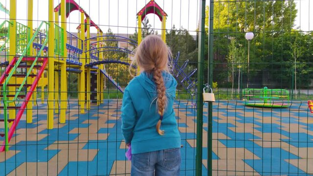 Sad lonely child near the closed playground because of the quarantine.A girl with long blonde braided hair staying alone near empty fenced showground without children.COVID-19 lockdown social distance