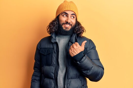 Young arab man wearing casual winter clothes smiling with happy face looking and pointing to the side with thumb up.