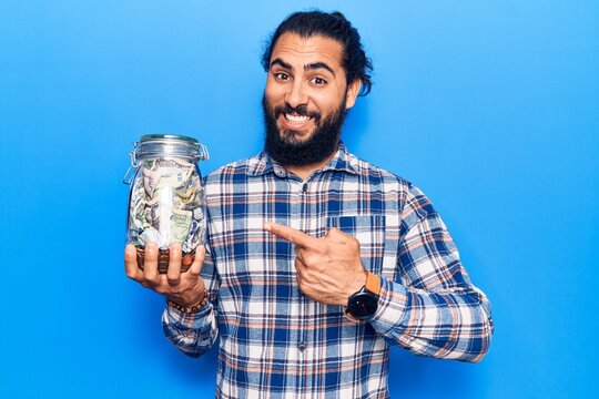 Young arab man holding jar with savings smiling happy pointing with hand and finger
