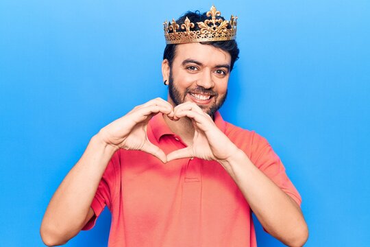 Young hispanic man wearing king crown smiling in love doing heart symbol shape with hands. romantic concept.