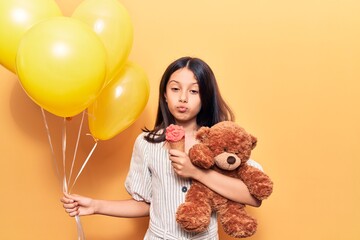 Adorable hispanic child girl holding balloons and teddy bear. Eating ice cream standing over isolated yellow background