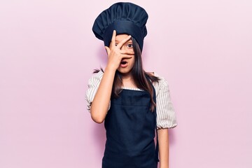 Beautiful child girl wearing cooker uniform peeking in shock covering face and eyes with hand, looking through fingers afraid