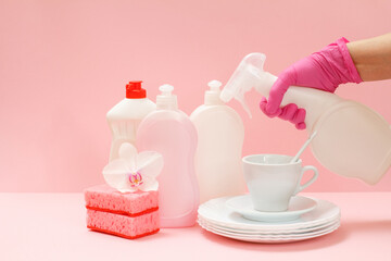 Woman's hand in nitrile glove with bottle of washing liquid and utensil on a pink background.