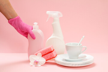 Woman's hand in nitrile glove with bottle of dishwashing liquid and utensil on a pink background.