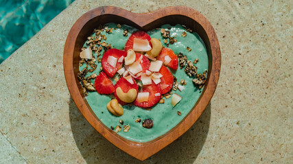 Green smoothie in a bowl in the form of a heart on the background of the pool on a bright sunny day