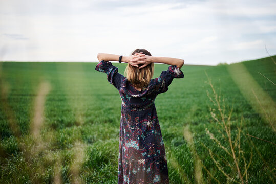 Young blond woman, wearing long dark boho dress, holding black hat standing with back to camera in green field in spring. Model posing outside in meadow. Hippie musician at natural environment.