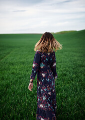 Young blond woman, wearing long dark boho dress, holding black hat walking away in green field in spring. Model posing outside in meadow. Hippie musician at natural environment.
