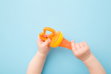 Infant hands holding orange nibbler on light blue table background. Baby accessory for chewing fresh fruit or berry through net sack of feeder. Pastel color. Closeup. Top down view.