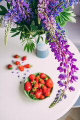 Obraz na płótnie Canvas A bouquet of lupins on a round table with strawberries. Bouquet of purple lupins in a grey-white-blue vase on a white roundtable with ripe strawberries in a green bowl, top view
