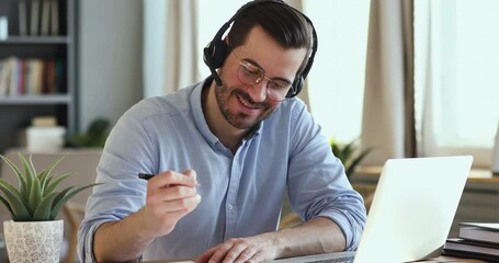 Focused young businessman in eyeglasses holding video conference call with clients, wearing wireless headphones with microphone. Skilled happy salesman advertising goods to customers, writing notes.