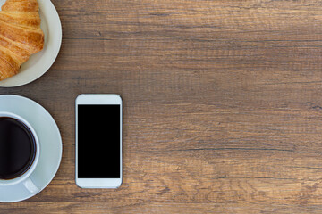 Top view of a desk with mobile phone, a cup of coffee and croissants on a wooden table.