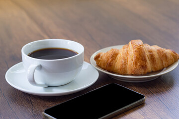 Side view of a desk with mobile phone, a cup of coffee and croissants on a wooden table.