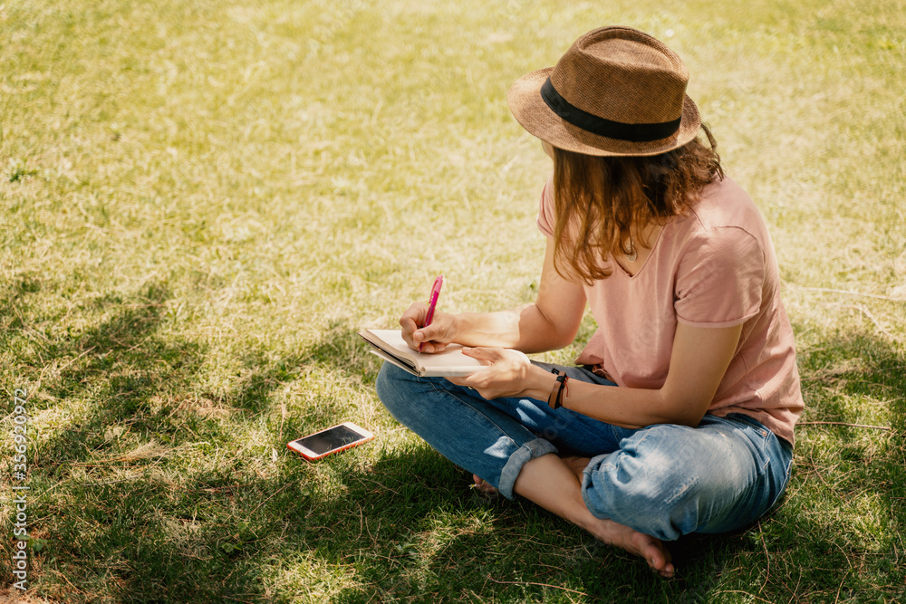 Wall mural summer sunny day. young woman sits in hat on a lawn and makes notes in a notebook. the girl works, s