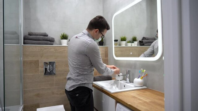 Young Caucasian guy in gray plaid shirt in disposable turquoise protective face mask wear black glasses opens tap, washes his hands, rubs with towel in nice bathroom in the hotel. Virus prevention.