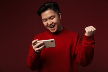 Image of excited asian man making winner gesture while using cellphone