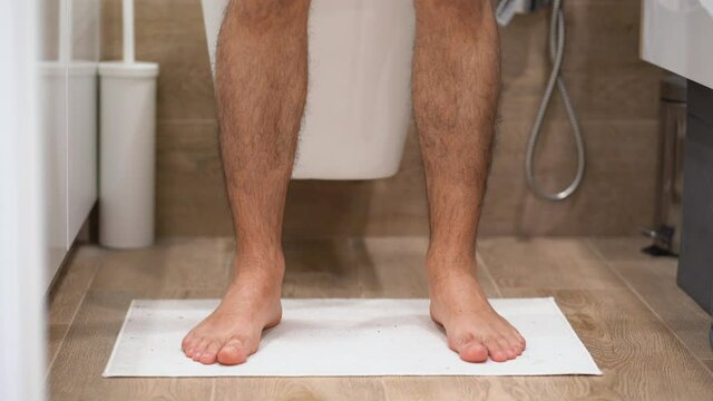 Young Caucasian thin guy in shirt with hairy legs enters bathroom, stand on white soft rug, sit down on toilet bowl, put off his gray underwear, office worker. Close up view of morning routine at home