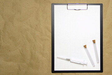 The tablet with a clip for the clip of paper with a white sheet a4 lies against the background of craft brown crumpled paper. On top is a medical syringe and two transparent empty tubes with stoppers.