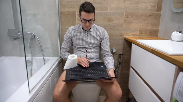 Young Caucasian pretty brunette entrepreneur man in black glasses, gray plaid shirt sit on white toilet bowl with underwear down, use laptop, surf. Businessman remote work at home in quarantine period