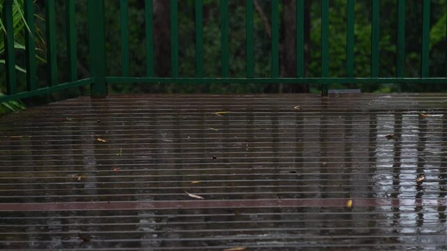 Dark brown wooden deck with green railing under a heavy rain. Rain drops with lots of water splashes. Green forest in background.