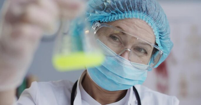 Close up of female Caucasian epidemiologist in mask, goggles and gloves holding glass flask with yellow liquid vaccine against virus in hands. Laboratory testing of vaccine. Clinic research concept