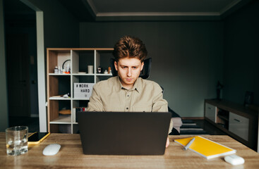 Freelancer in a shirt roasting at home at a table on a laptop online, working online during quarantine. Young male programmer working at home as a freelancer. Remote work.