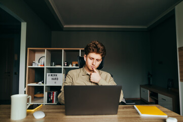 Pensive serious freelancer in a shirt and wireless headphones works remotely from home, looking at the screen with a focused face. Business young man working at home teleworking.