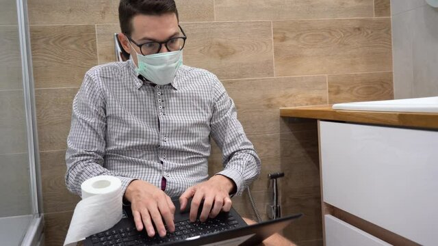 Young serious man wear gray plaid shirt, black glasses, protective medical face mask sits on toilet bowl and uses laptop. Business guy remote working at home in bathroom during quarantine by covid-19