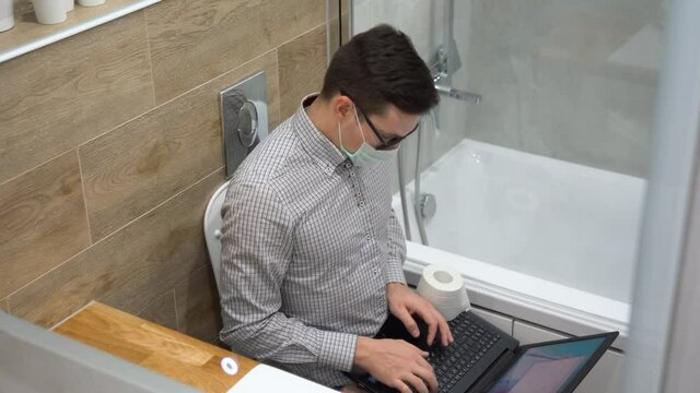 Young Caucasian brunette entrepreneur man in black glasses, gray plaid shirt, face mask sit on white toilet bowl, use laptop, surf internet, make job in lavatory. Remote home work in quarantine time