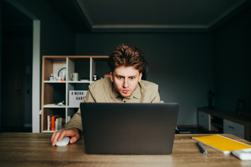Emotional young man in a shirt working at home on a laptop with a tense face, looking at the screen. Dissatisfied freelancer works at work at home, uses a laptop with an evil face.