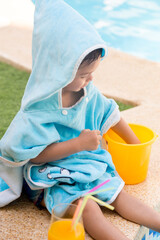 Child sitting with a hooded towel playing with a plastic bucket and an orange juice in a pool