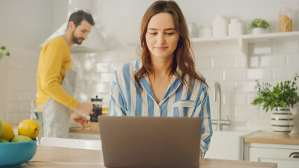 Obraz premium Young Beautiful Woman Using Laptop Computer While Wearing Blue Pyjamas. Brunette Female Sitting in a Modern Sunny Kitchen Room. Freelancer Working from Home.