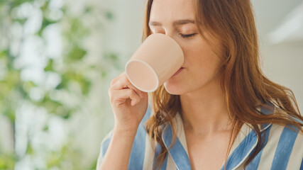 Authentic Portrait of a Beautiful Young Female in a Bright Room at Home Drinking Tea or Coffee from a Cup. Attractive Brunette Woman Happy Morning Routine.