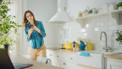 Attractive Young Female in Jeans Shirt and Brown Pants is Using Her Smartphone in the Kitchen. She Dances in a Modern Kitchen with Healthy Lifestyle Vibes. Happy Beautiful Girl Relaxing at Home.