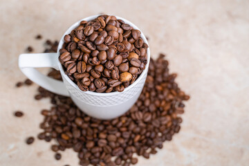 a white cup filled with roasted cinnamon coffee beans standing on brown marble surrounded by coffee beans