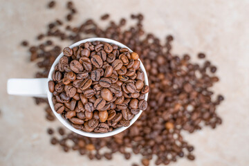 a white cup filled with roasted cinnamon coffee beans standing on brown marble surrounded by coffee beans