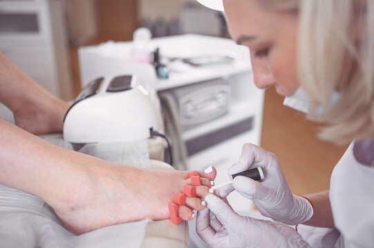 Pedicurist doing white nail polish on client legs using shellac lamp. Professional medical pedicure procedure. Foot treatment in SPA salon. Podiatry clinic. Beautician doctor hands in white gloves.