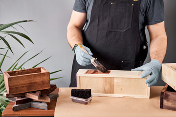Man carpenter varnishing wooden crate for flowers with brush in her small business woodwork workshop. In your work, do you use stains or wood preservatives to show the wood pattern.